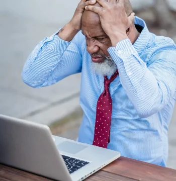 Senior anxious black businessman in elegant blue shirt and tie grabbing head while sitting at wooden table with laptop outdoors
